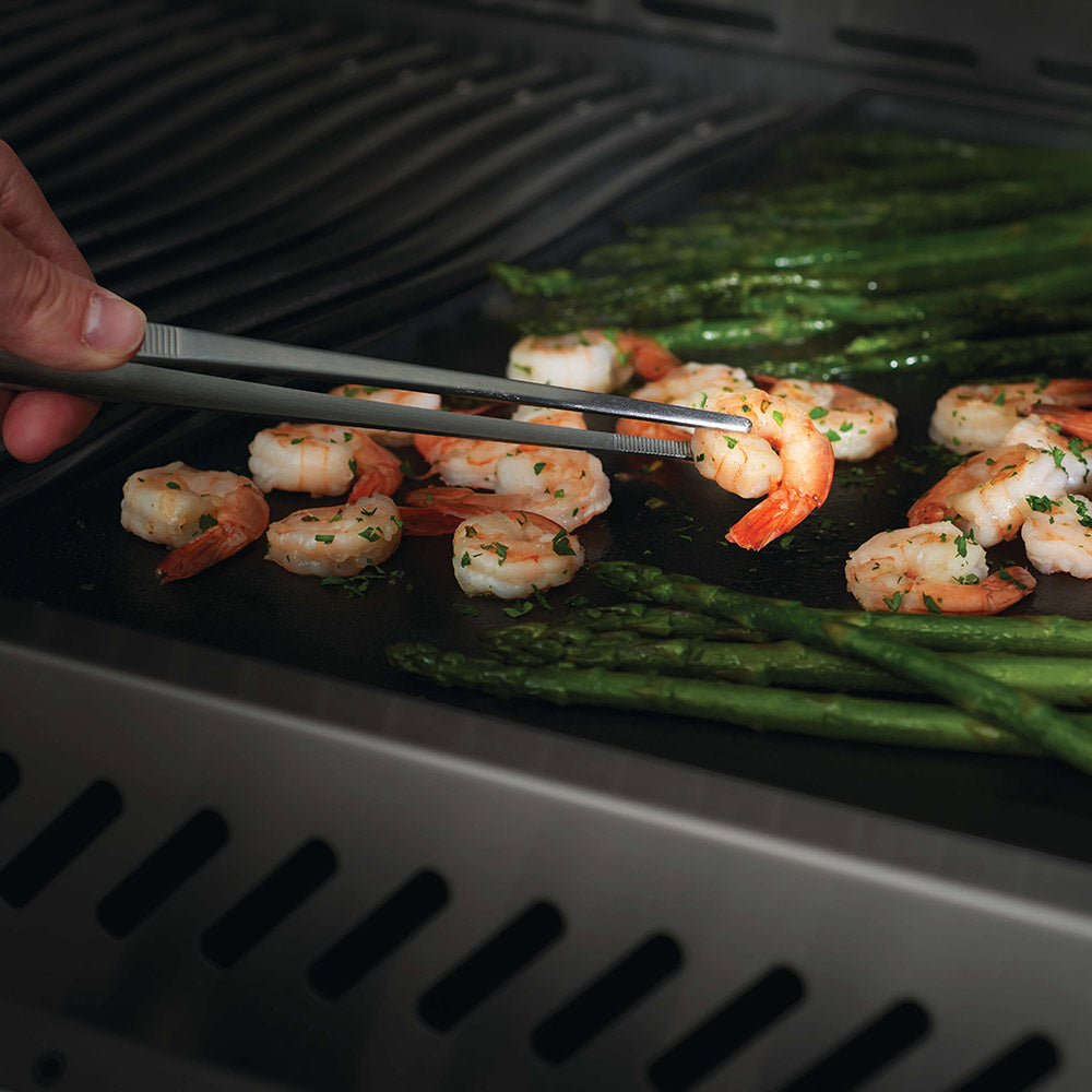 Stainless steel grilling tongs gripping shrimp and asparagus on a hot grill grate