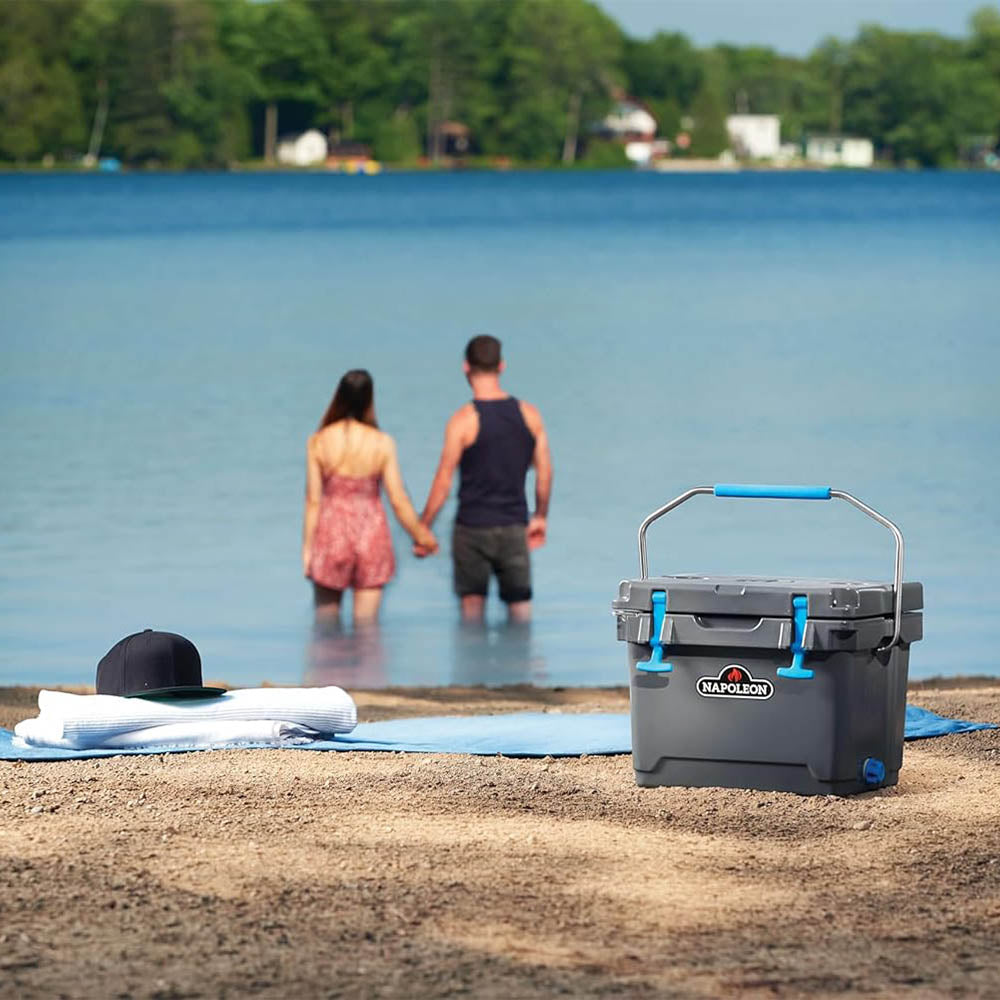 Gray cooler chest with blue handle and Napoleon logo, closed lid on beach