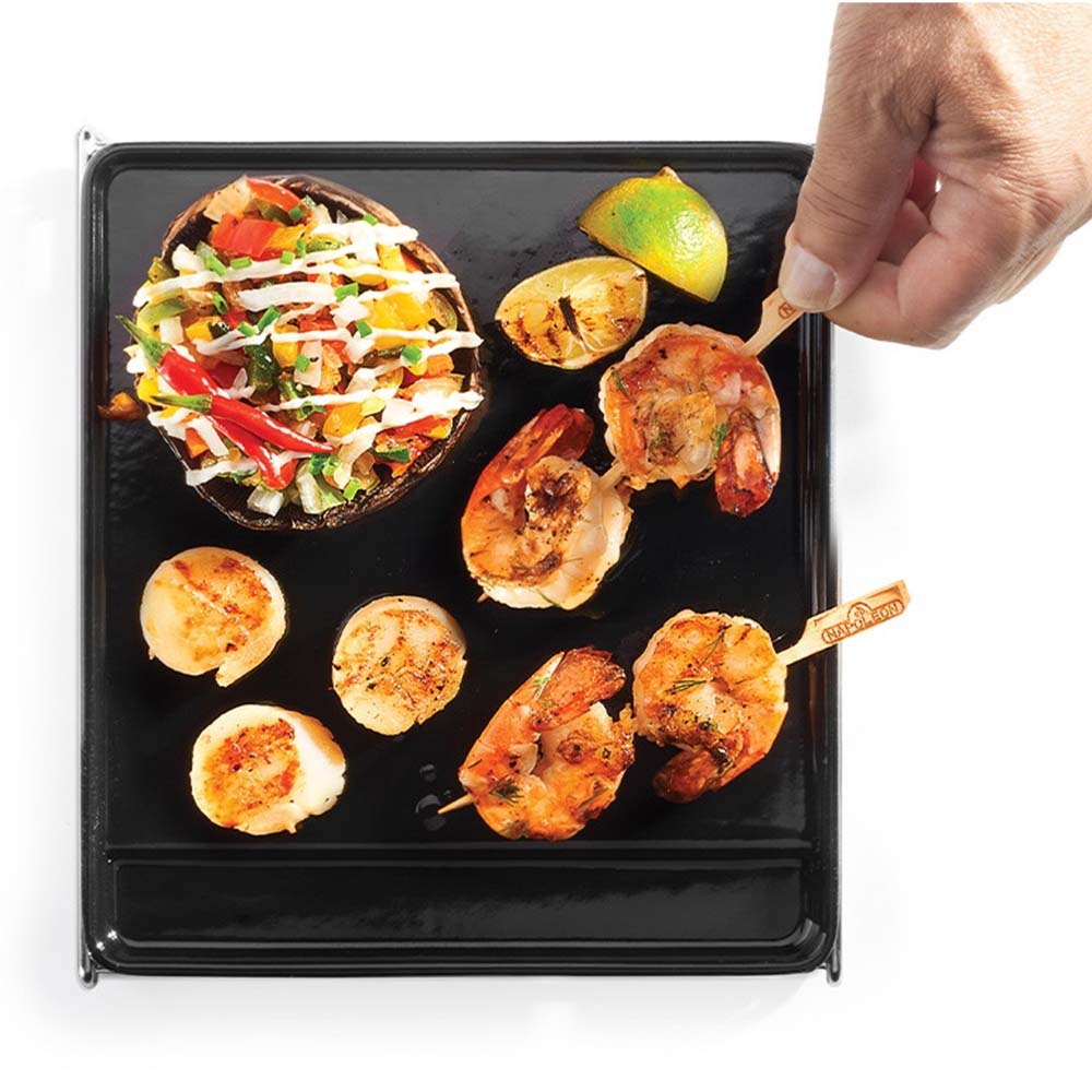 Cast iron griddle cooking shrimp and vegetables on black surface; hand holds skewer