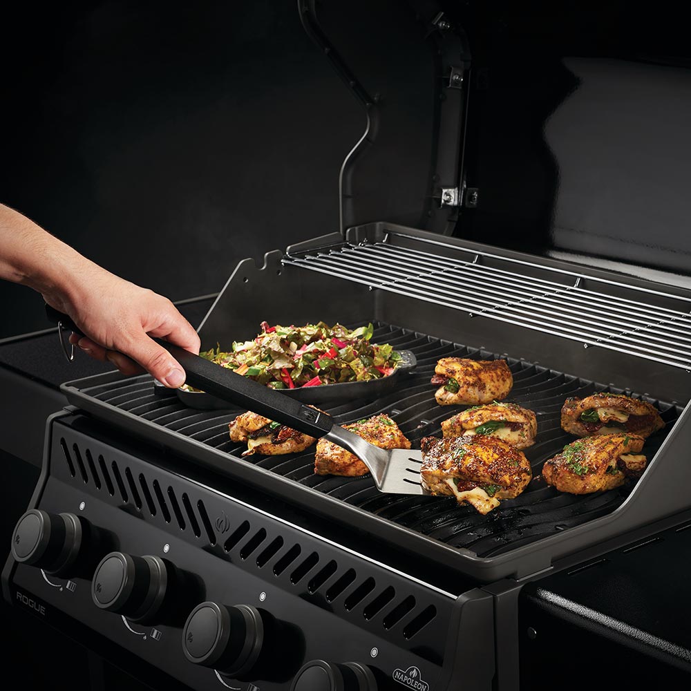 BBQ spatula arranging grilled chicken on the grates while grilling vegetables nearby
