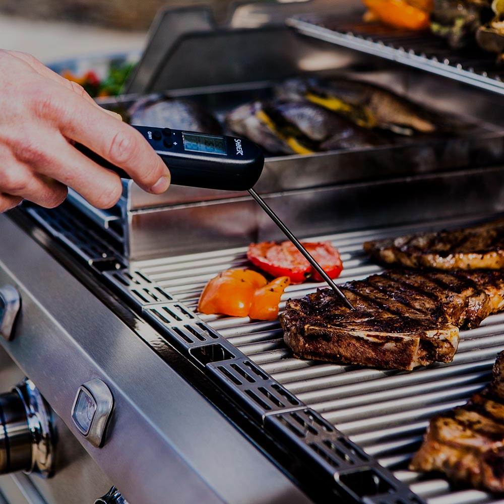 Saber EZ Temp digital thermometer held above grill, metal probe in food