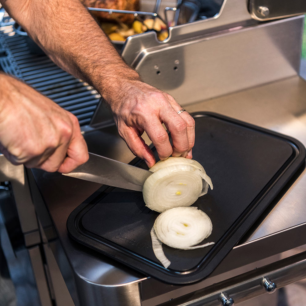 Black roasting pan on grill with onion slices being placed by hands