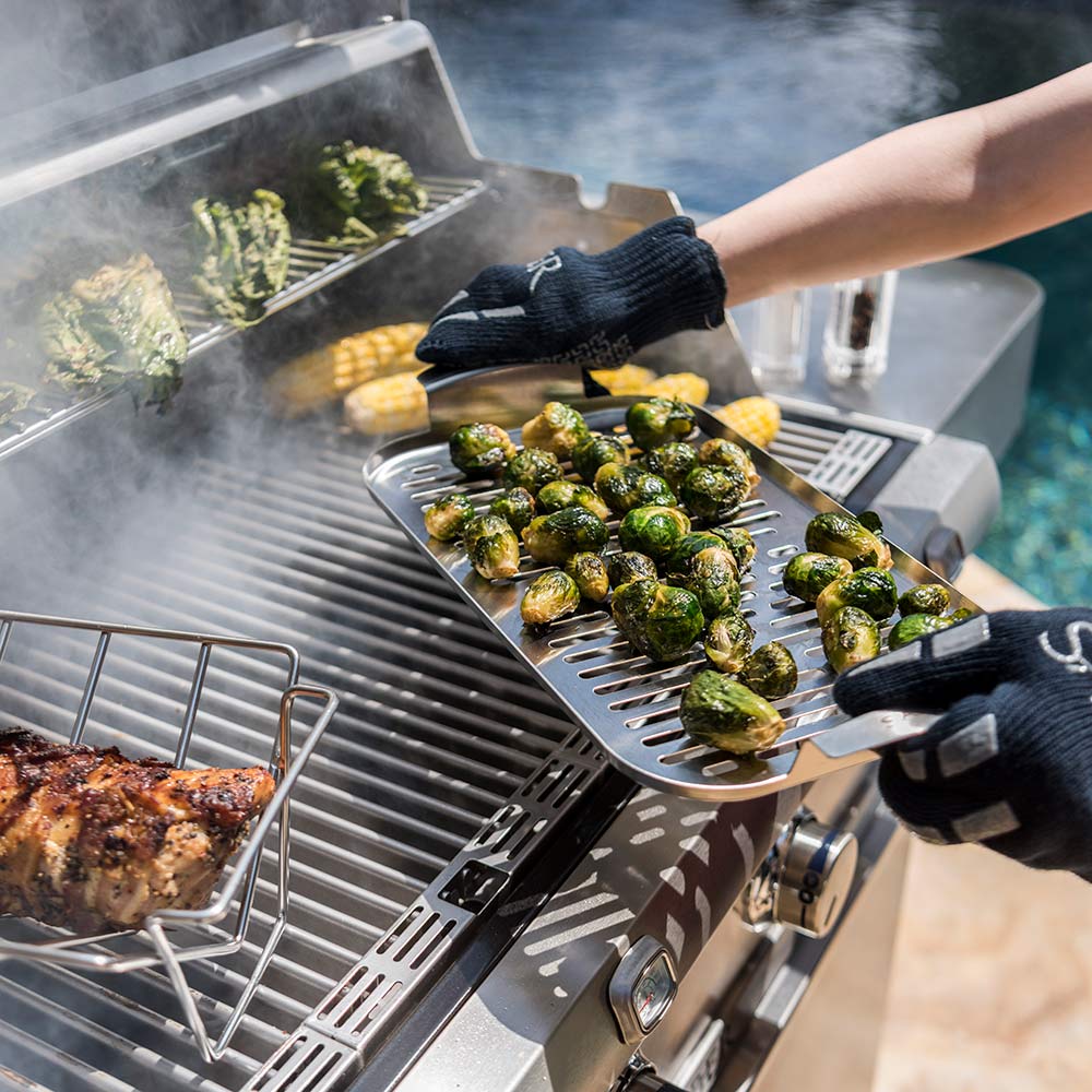 Stainless steel steamer tray with vegetables being set onto a grill