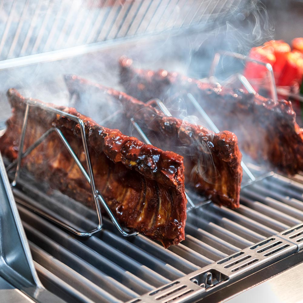 Stainless steel rib rack holding grilled meat over grill grates with visible smoke