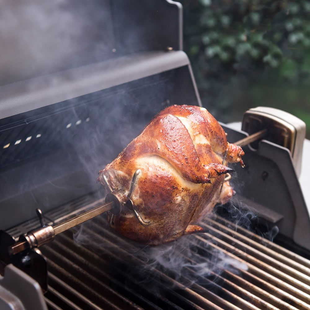 Rotisserie chicken turning on stainless spit over grill grates