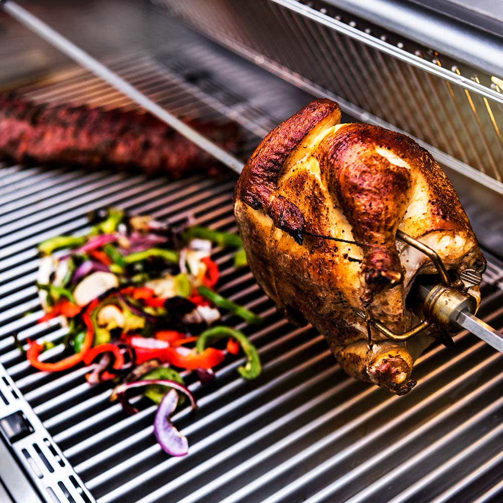 Rotisserie chicken on spit with vegetables on the drip tray inside grill