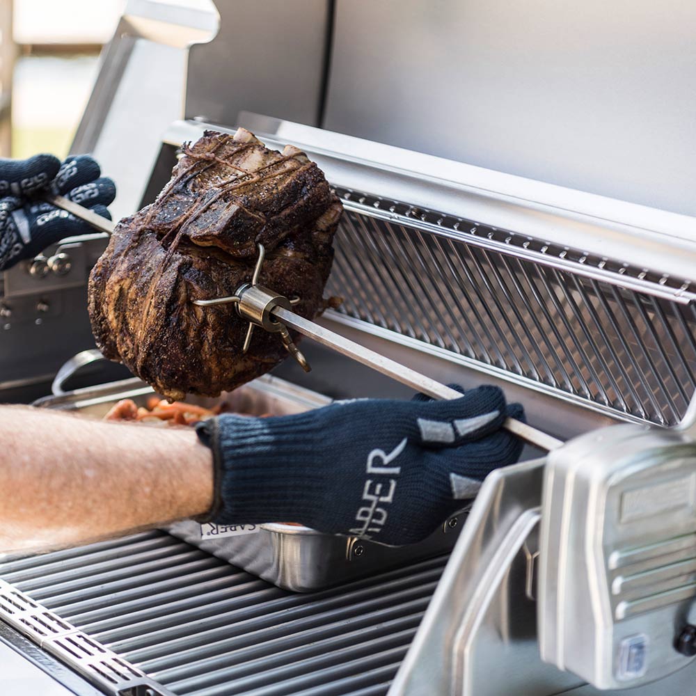 Rotisserie chicken on spit installed in grill with gloved hands adjusting the kit