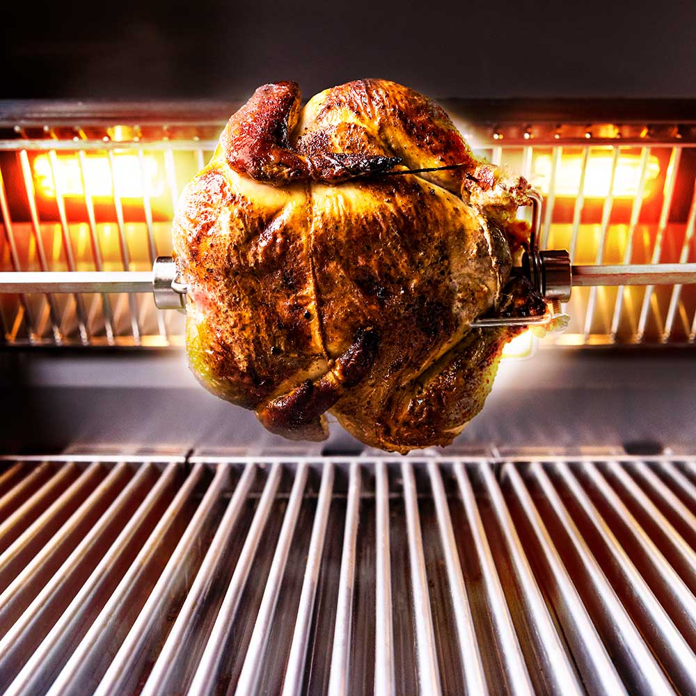 Rotisserie rod holding whole chicken rotating over grill flames