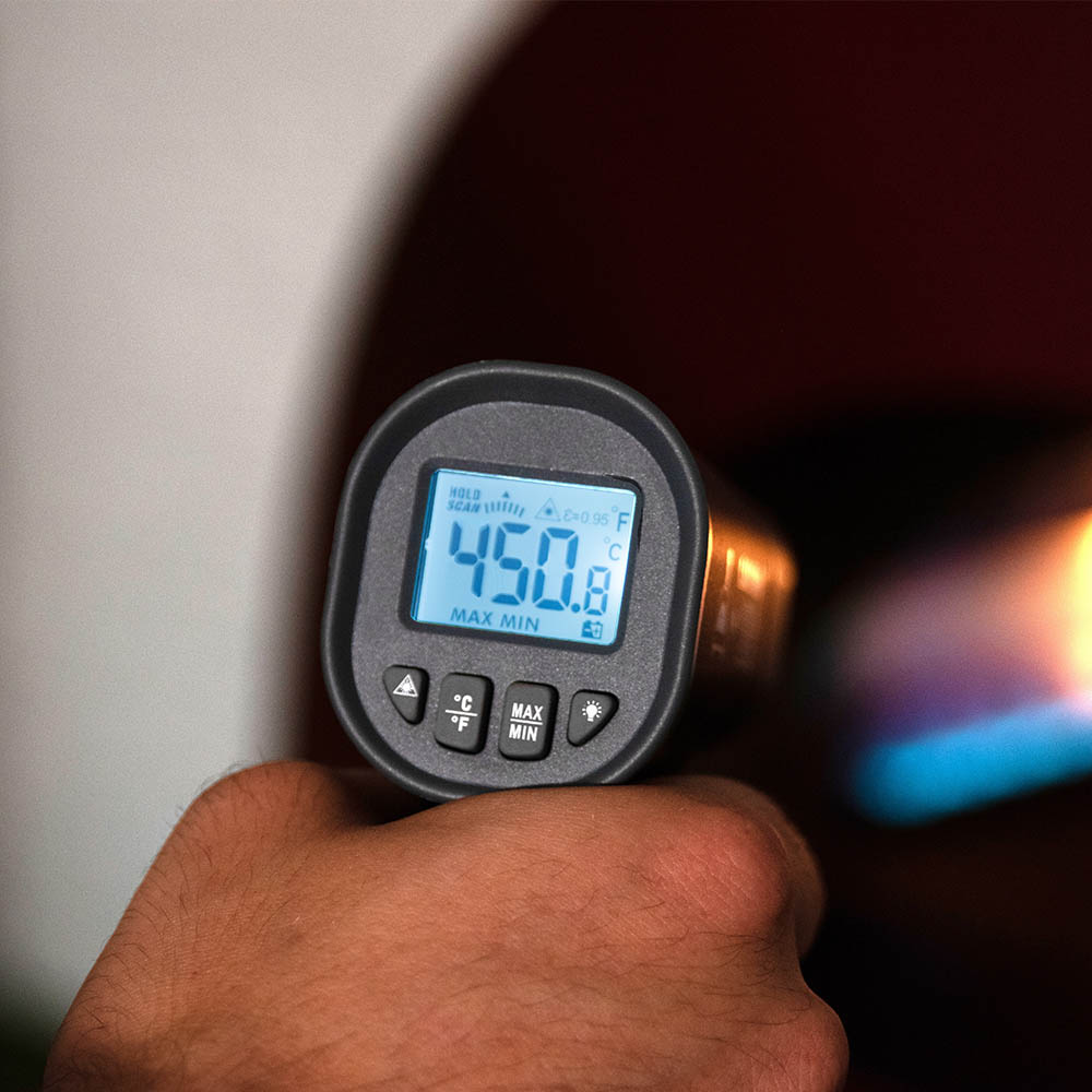 Handheld laser thermometer in use showing blue LCD temperature readout