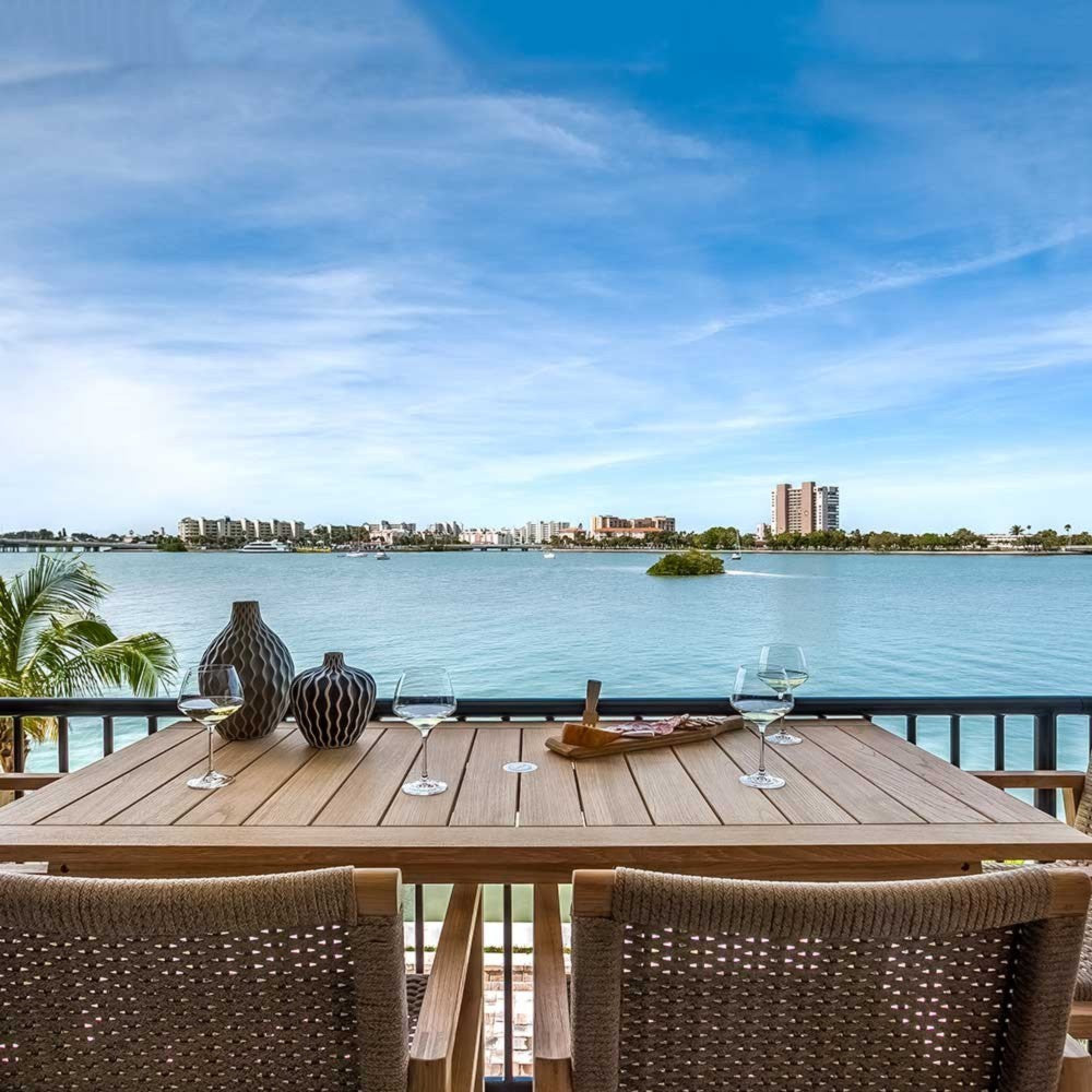 Outdoor counter-height teak dining table with woven chairs and rectangular tabletop by railing.