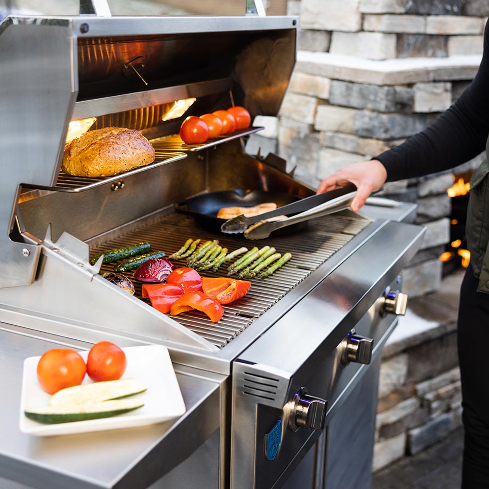Open stainless gas grill with vegetables and whole chicken on grates; hand holding tongs.