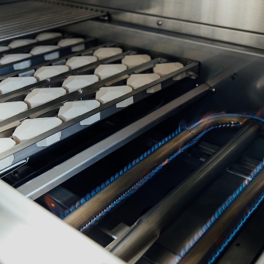 Gas grill burner tubes with blue flames inside stainless cooking chamber.