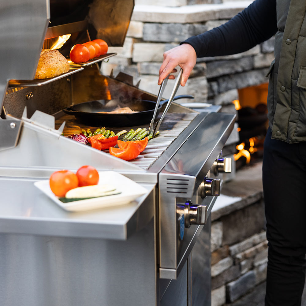 Gas grill with open hood and cooking grates, food in trays with tongs