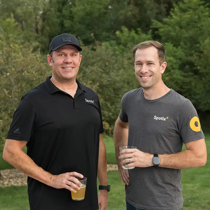 Two men standing outdoors, both wearing Spotix-branded clothing, holding drinks.