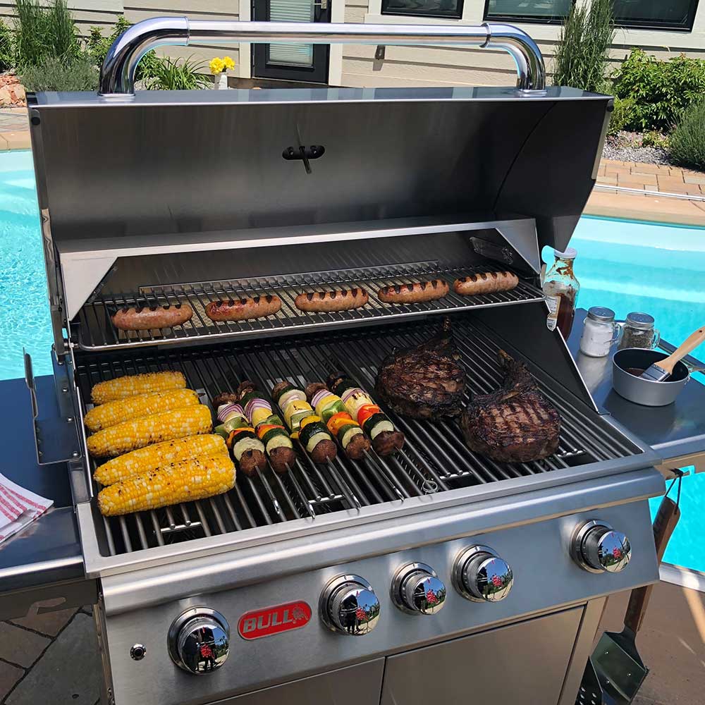 Open stainless steel gas grill with sausages, steaks, and corn on the grates