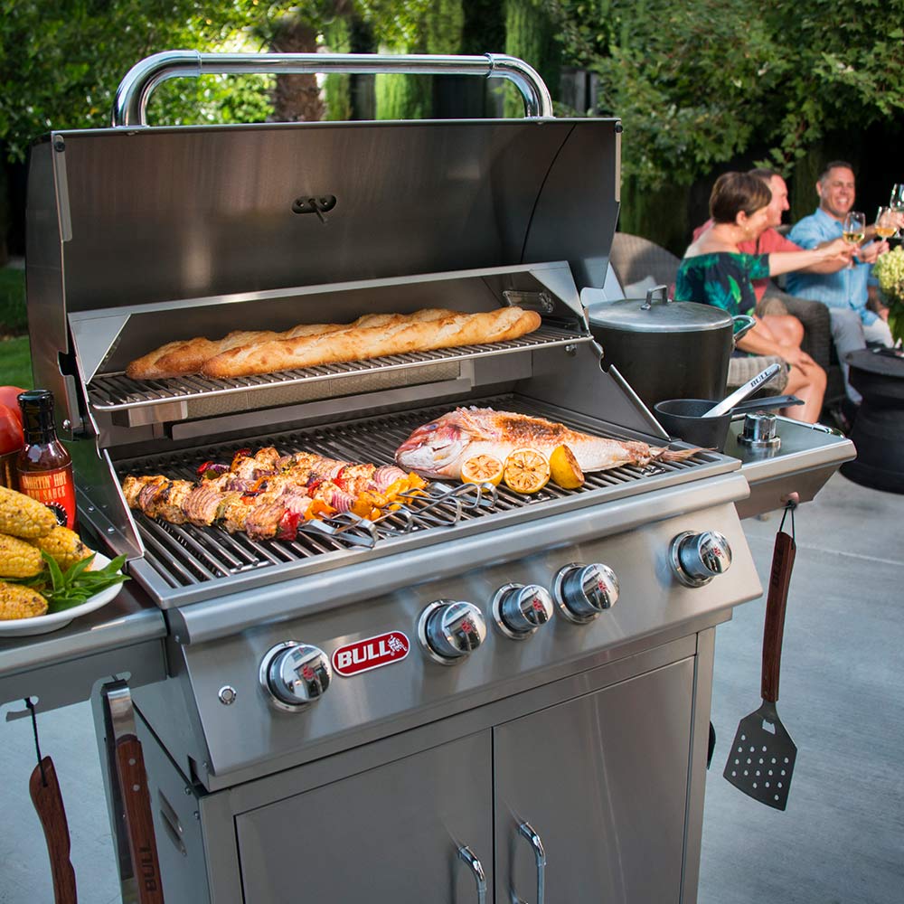 Open gas grill with multiple cooked items on grill grates and front control knobs