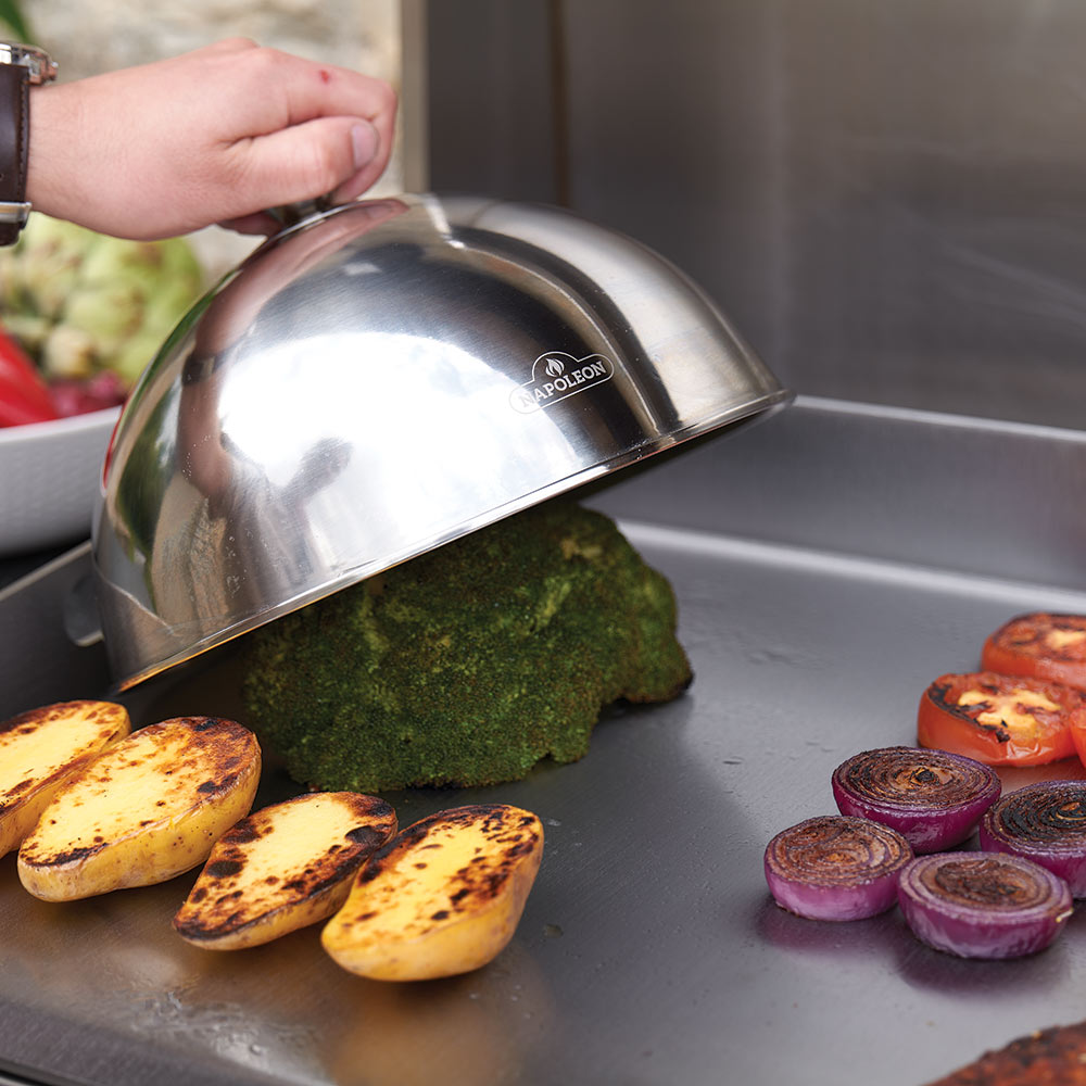 Flat top griddle with a smoking dome over greens, with grilled potatoes and onions