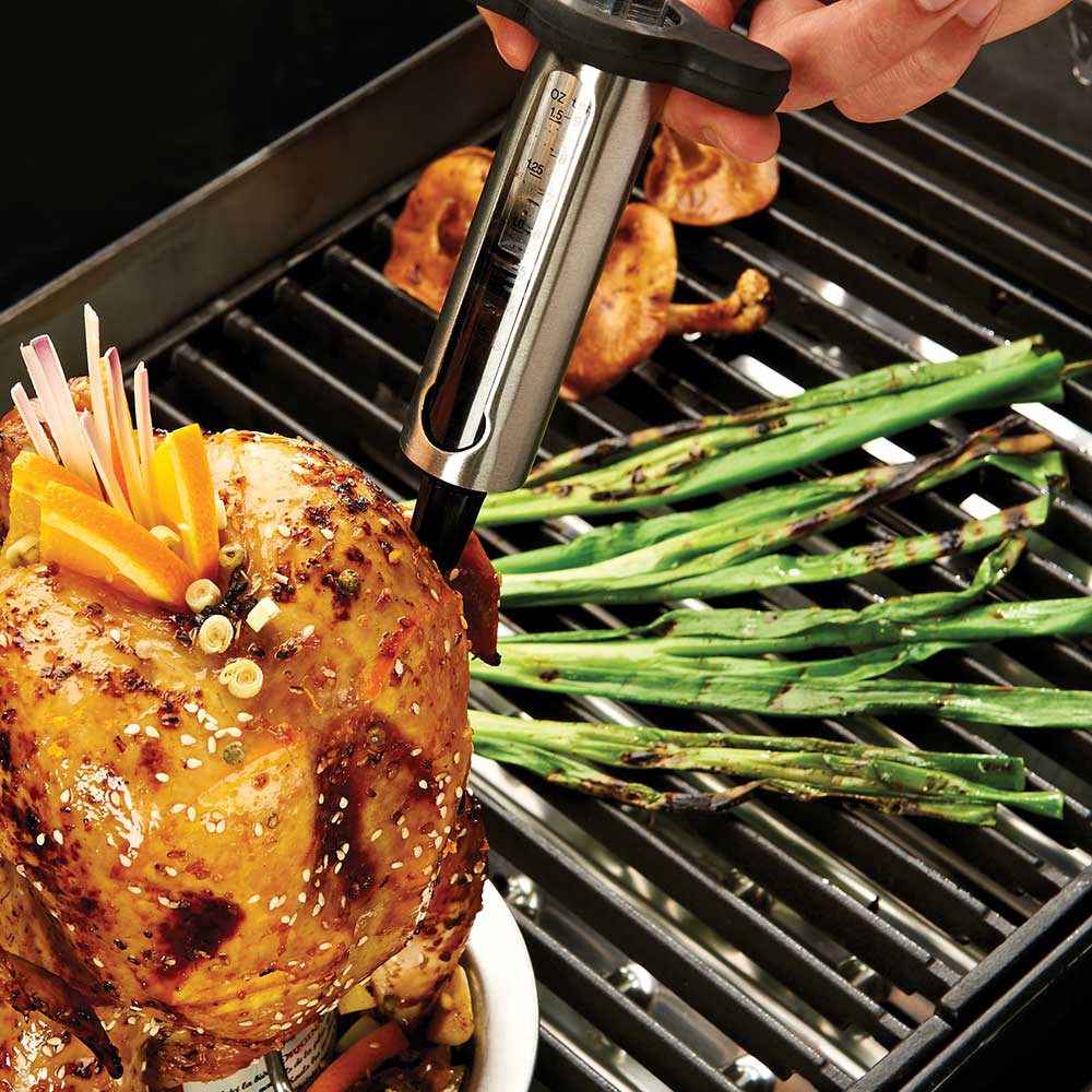Marinade injector inserted into chicken on the grill next to green onions