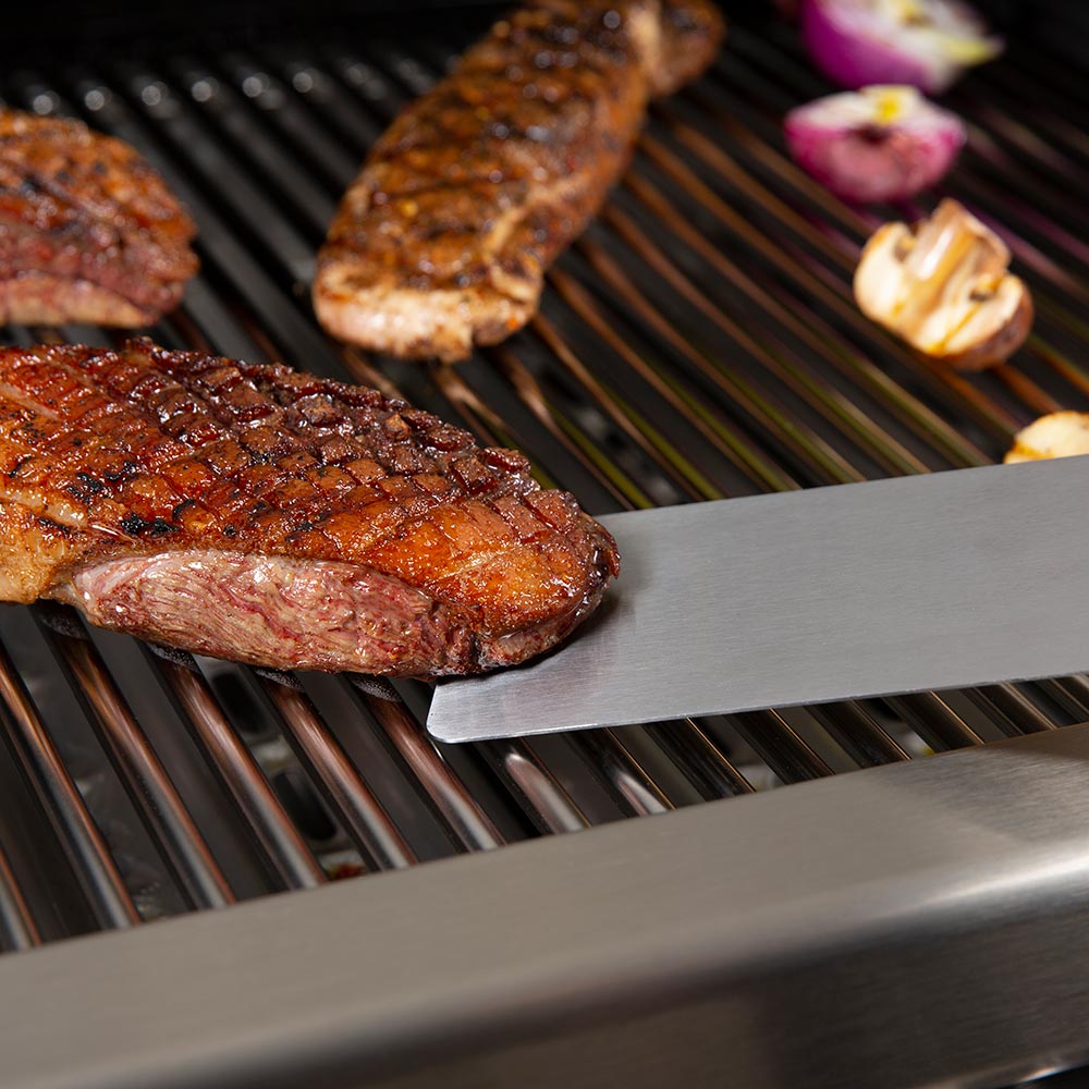 Angled stainless flipper blade with black handle holding a grilled steak