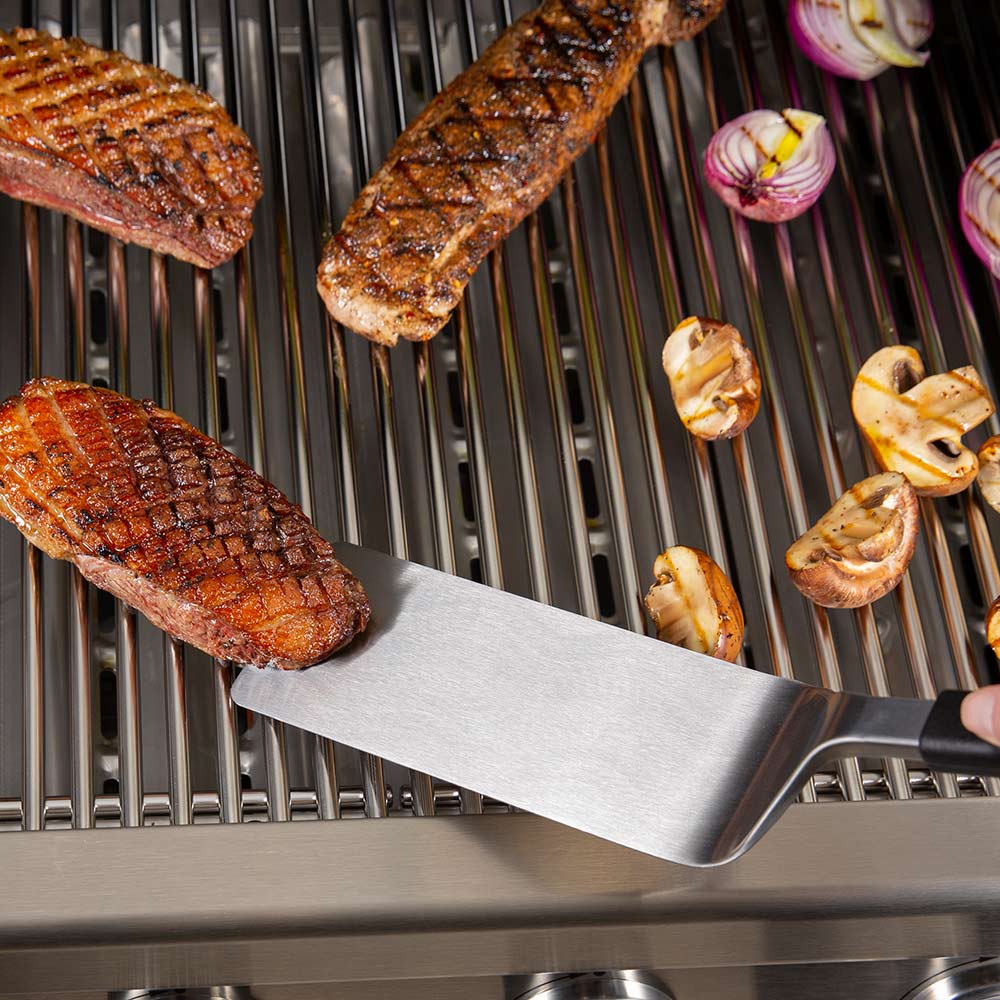 Grill flipper spatula with wide stainless blade and black handle on grill grates