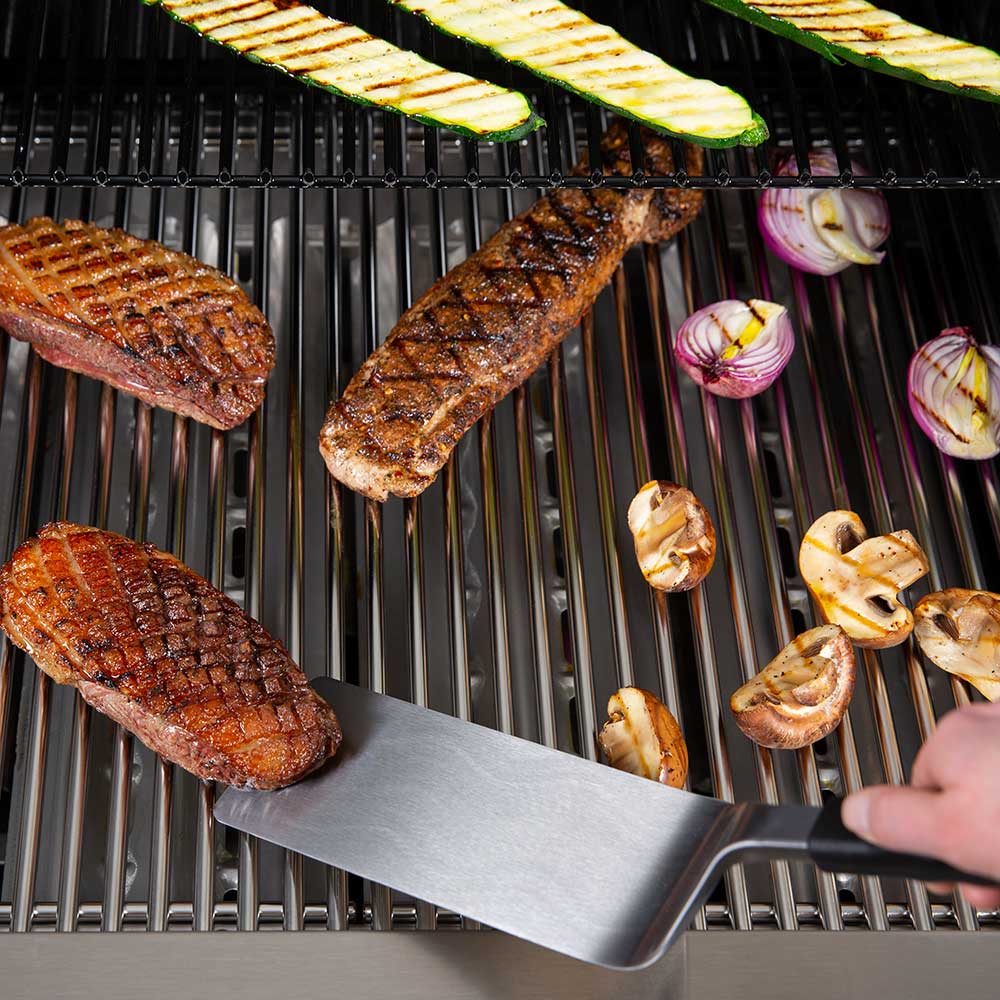 Wide stainless grill flipper blade with black handle resting on grill grates