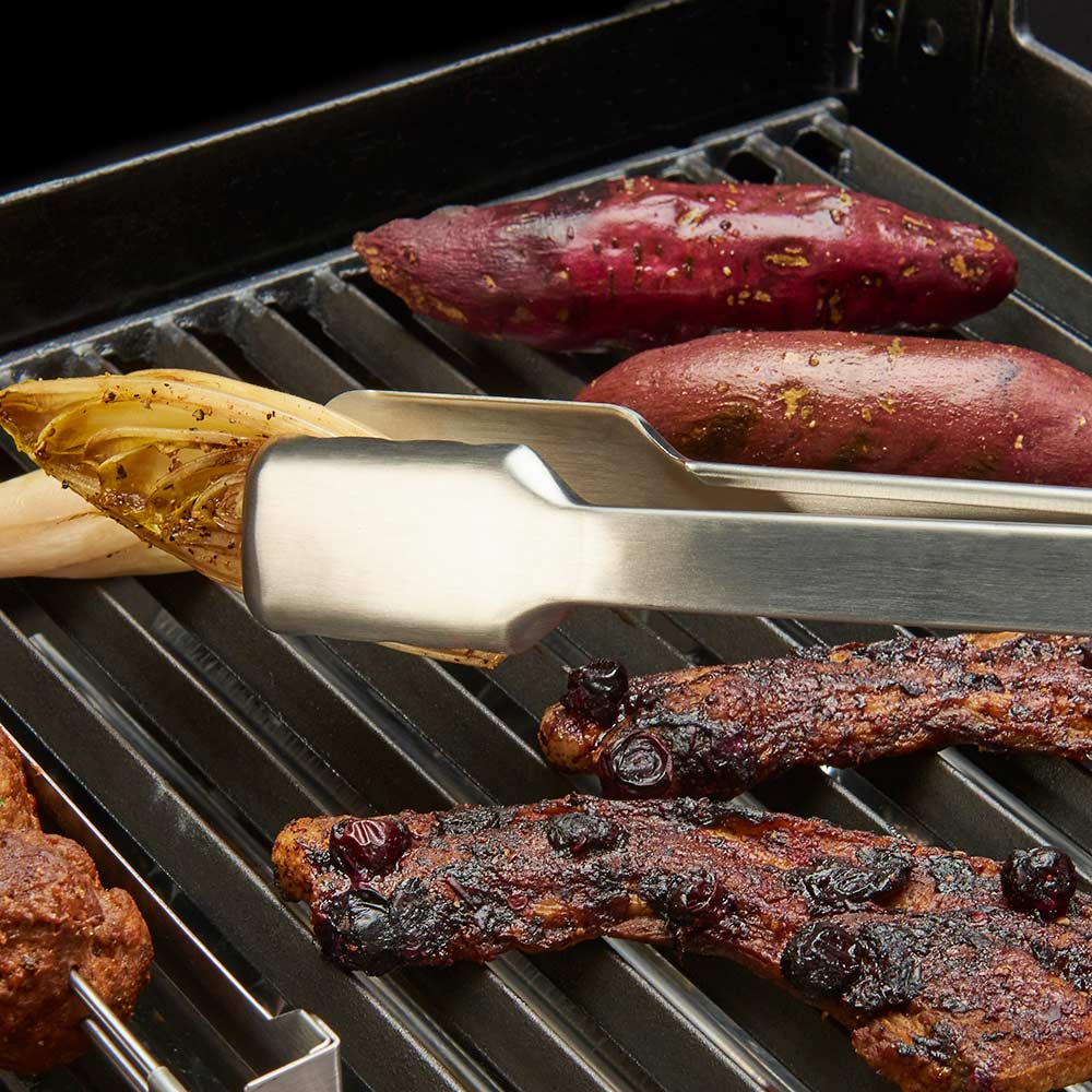 Stainless grill tongs gripping roasted food over grill grates.