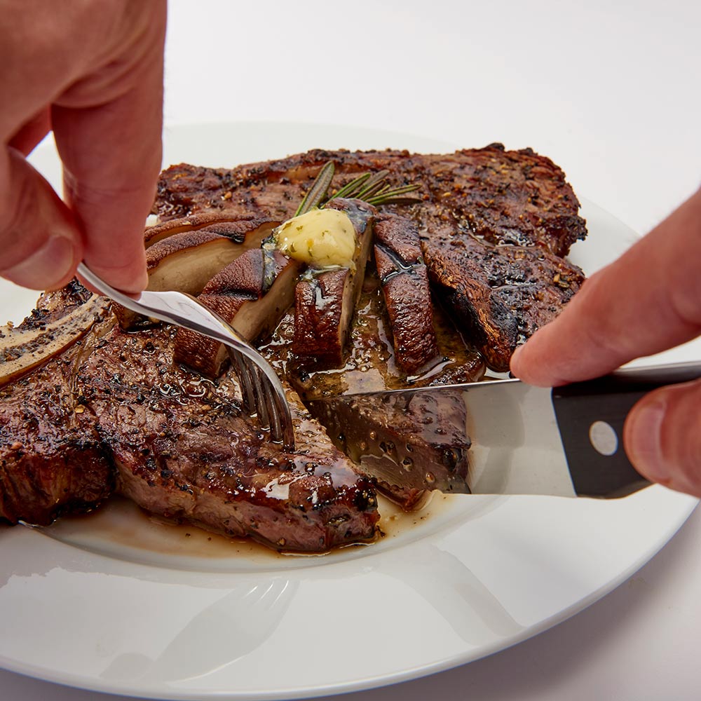 Hands using a steak knife on cooked steak, black handle and serrated blade visible