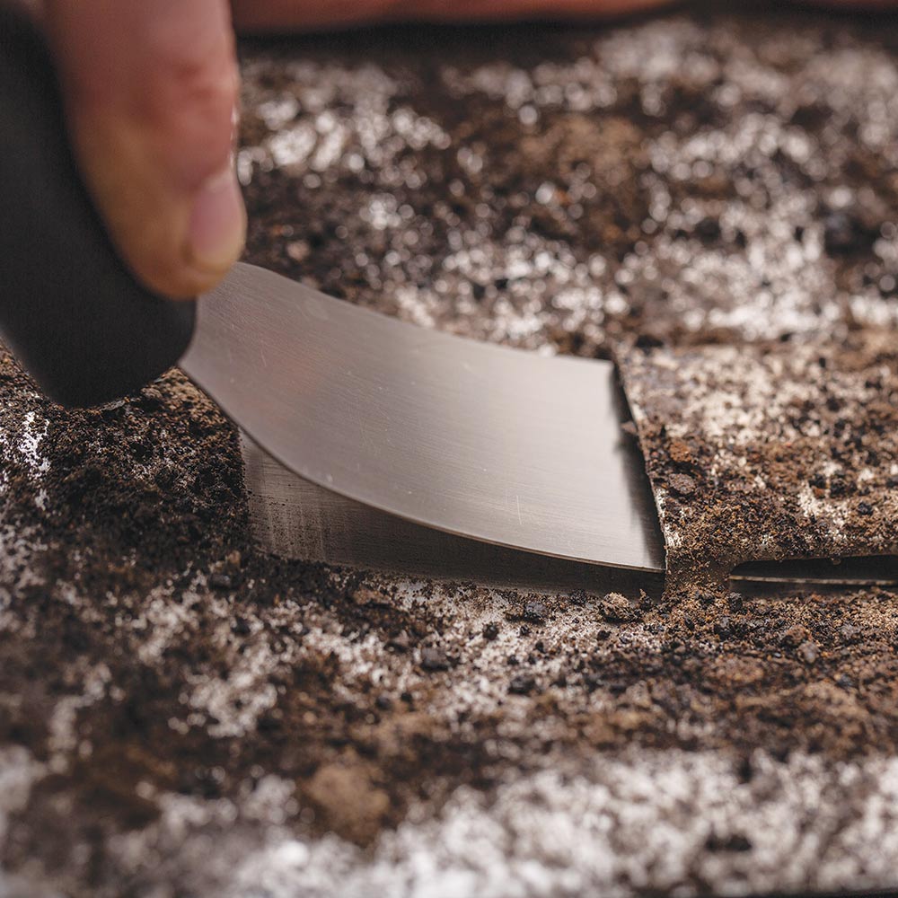 Metal scraper blade lifting char and ash from a grill surface