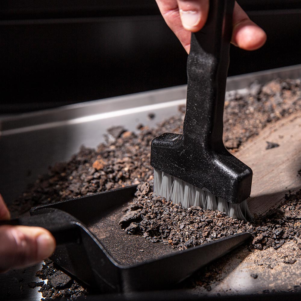 Black brush with white bristles scrubbing ash inside a grill tray
