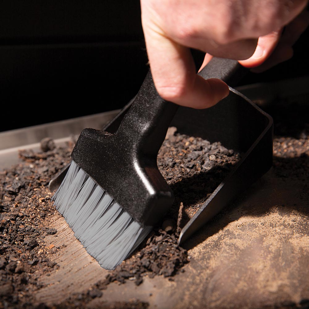 Black bristle brush sweeping ash into a grill cleaning pan