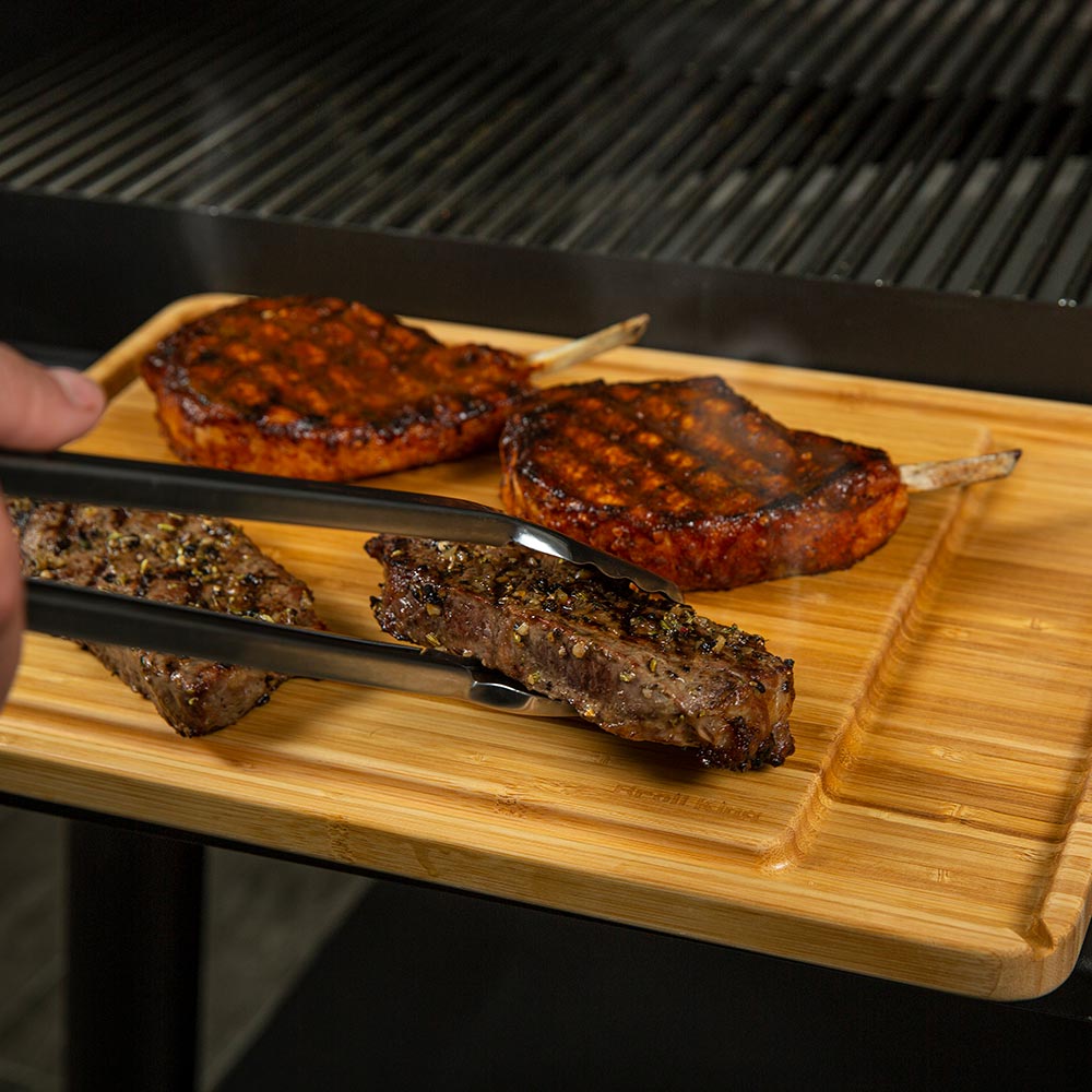 Bamboo serving board with grilled chops being lifted on black tongs