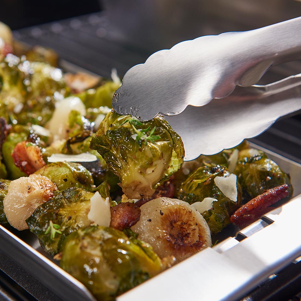 Narrow stainless steel grill topper tray with roasted vegetables, utensil lifting vegetables