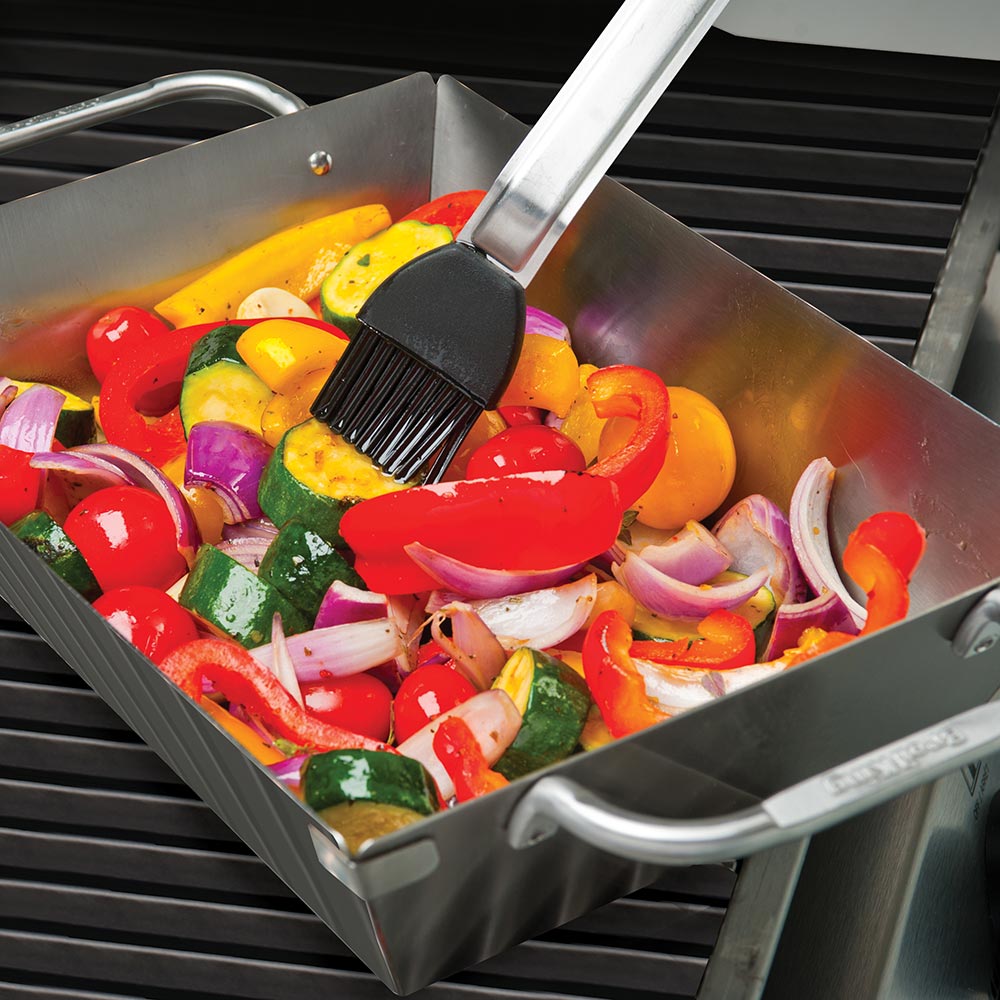 Stainless steel grill wok topper with side handles on grill, vegetables being sautéed.