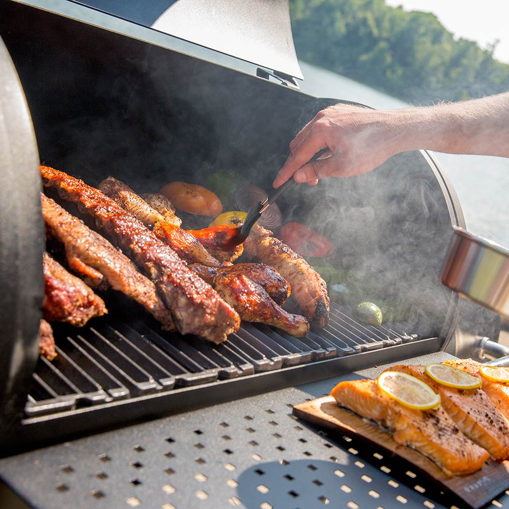 Charcoal smoker grill with skewered sausages and other meat over dark grates, smoke rising