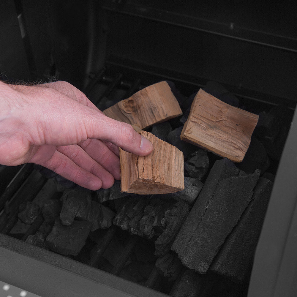 Hand placing wood chunks into the charcoal smoker firebox over dark briquettes