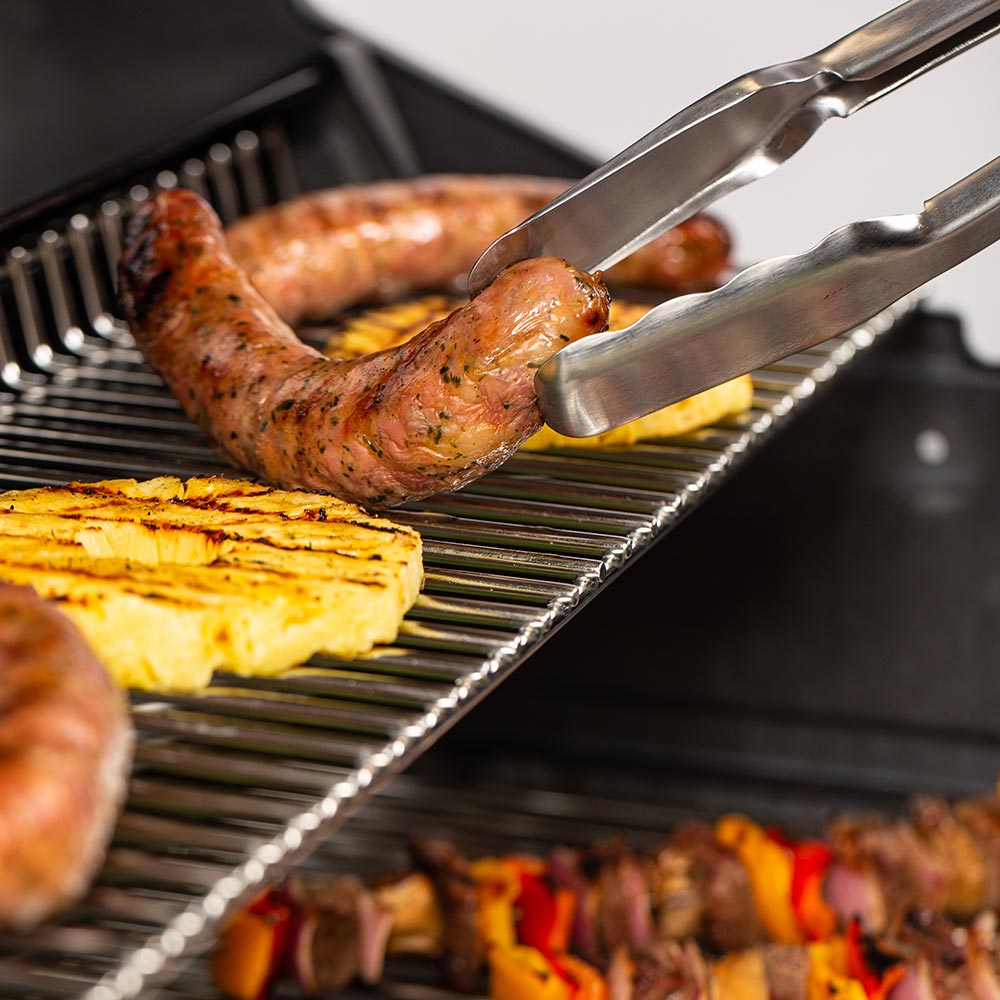 Grilled sausages and sliced food on stainless grill grates with metal tongs
