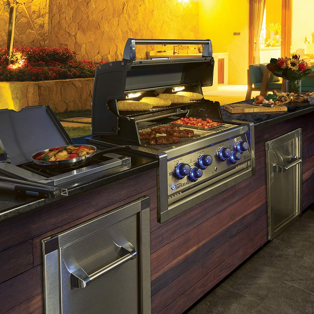 Built-in gas grill with open hood, lit burners, and side food prep surfaces