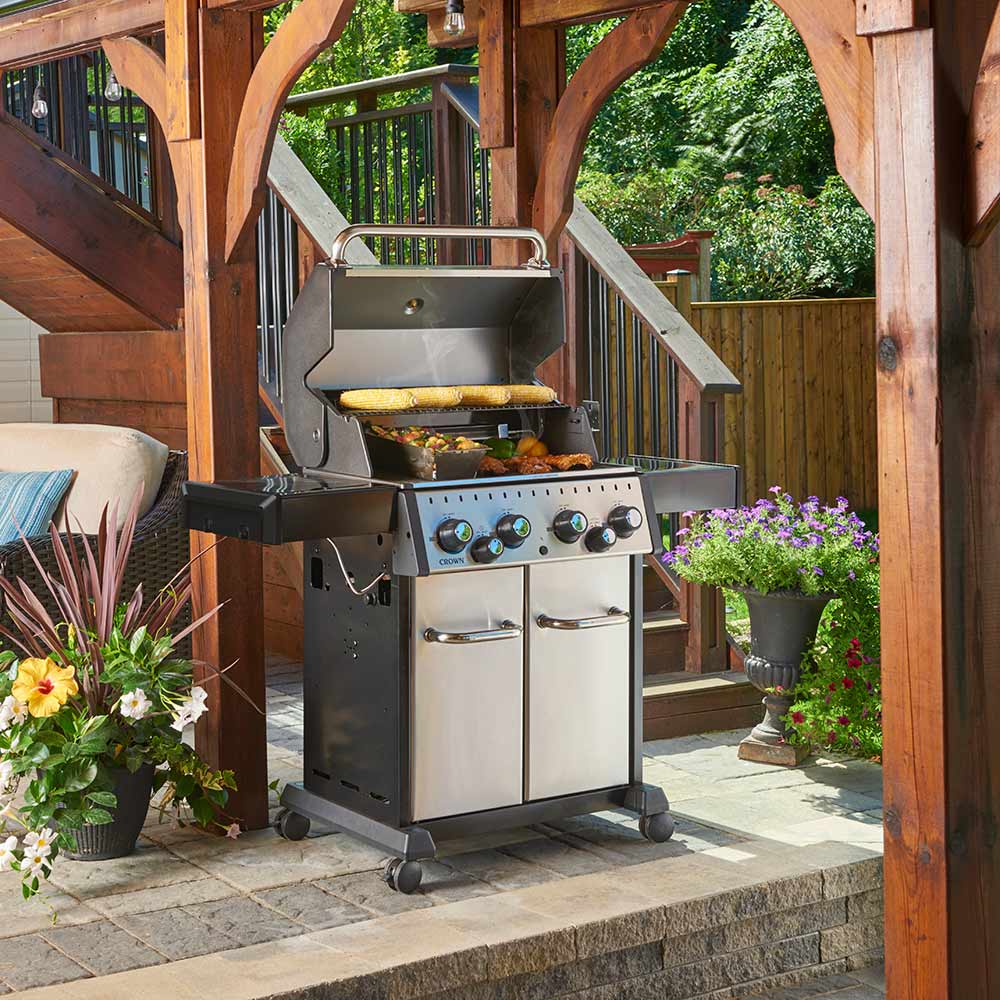 Stainless steel 4-burner gas grill with food on the cooking grate, lid open, patio view