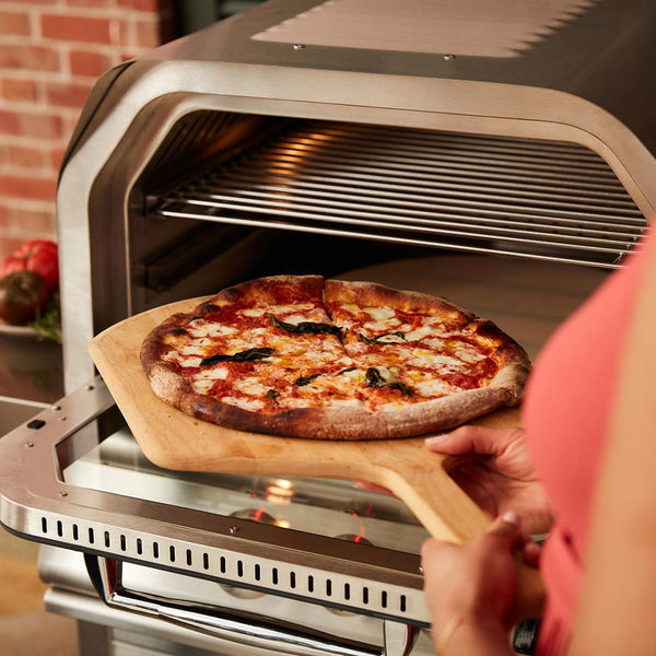 Cheese-topped pizza on wooden peel inside gas pizza oven with open front