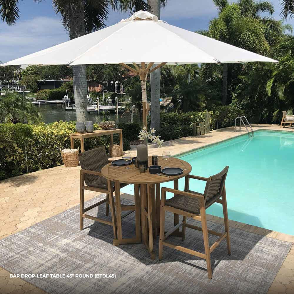 Round drop-leaf teak bar table with slatted top beside patio chairs and umbrella