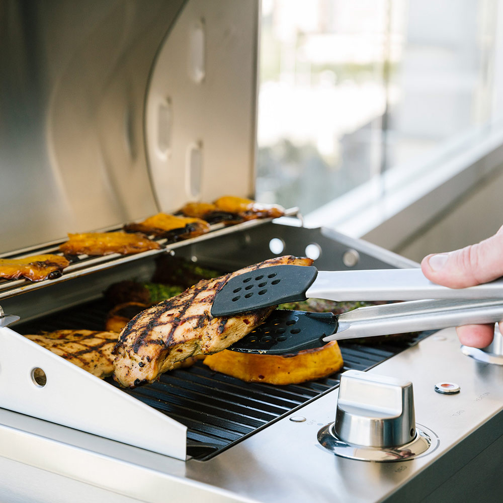 Stainless electric grill with steaks cooking on grates, spatulas nearby