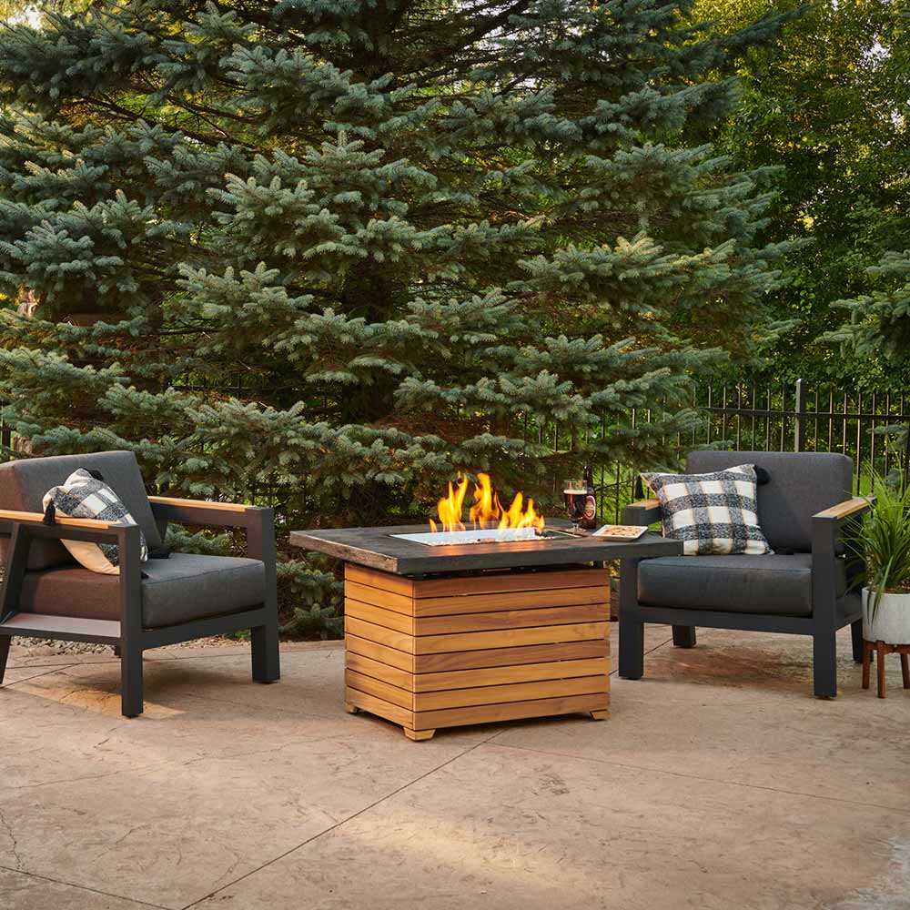 Outdoor gas fire pit table with glass firebox and flames, dark top and stacked teak base