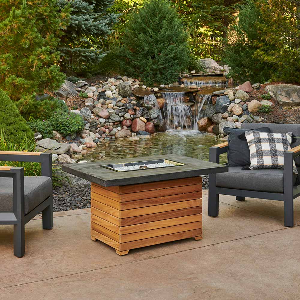 Outdoor gas fire pit table with glass firebox, flames visible, dark top and teak base