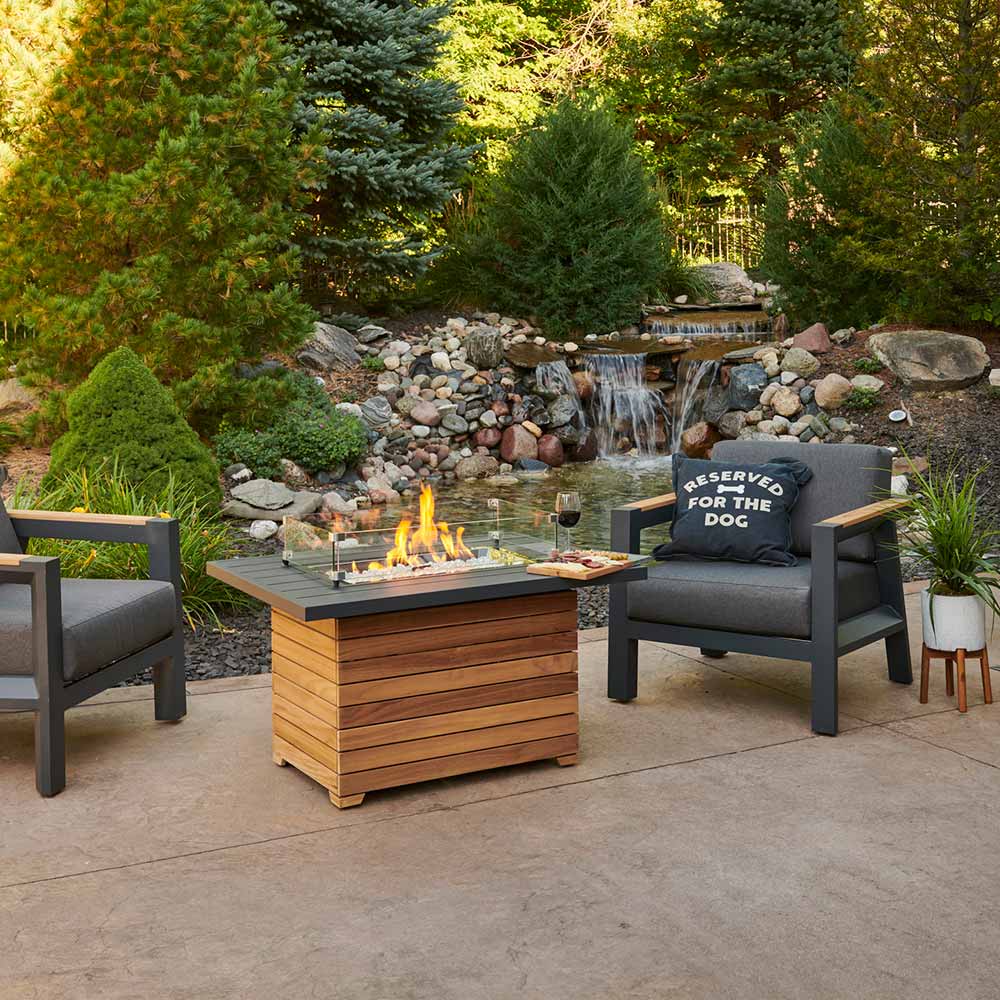 Outdoor gas fire pit table with dark aluminum top and teak base, flames and seating nearby