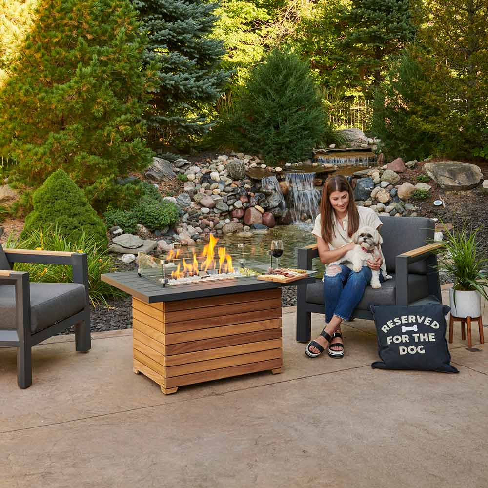 Outdoor gas fire pit table with dark aluminum top and teak base, flames in use