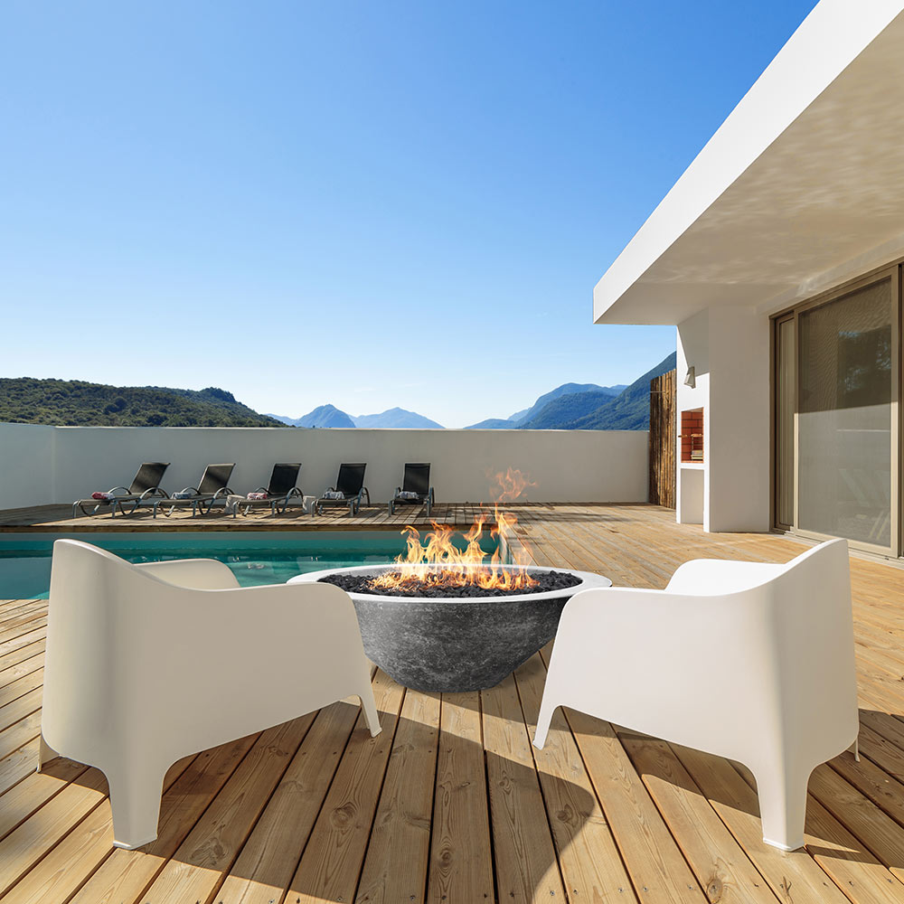 Concrete gas fire pit on deck with flame, set among outdoor dining chairs.