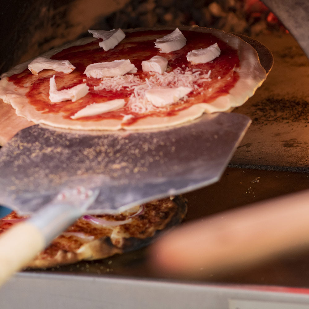 Aluminum pizza peel holding a pizza with white cheese and red sauce near oven