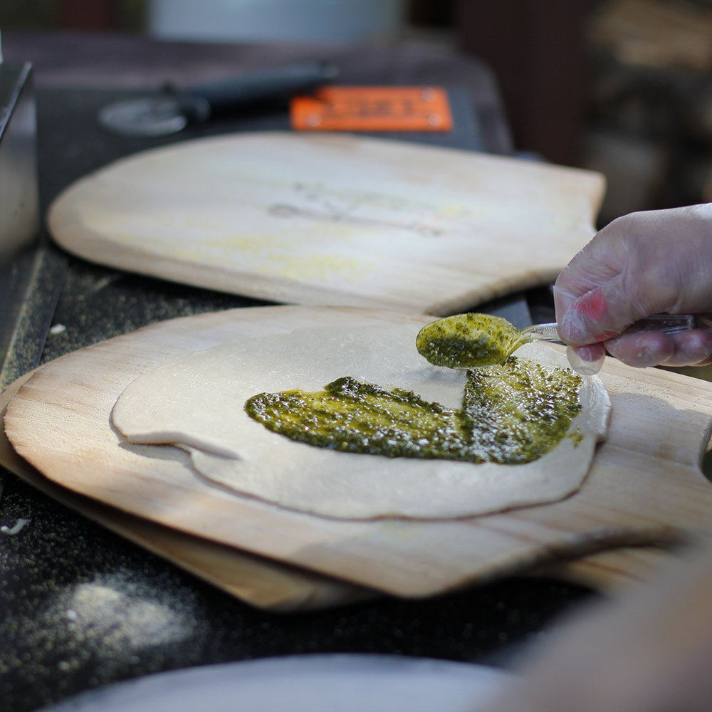 Wooden pizza paddle with dough topped with green pesto, hand spreading sauce