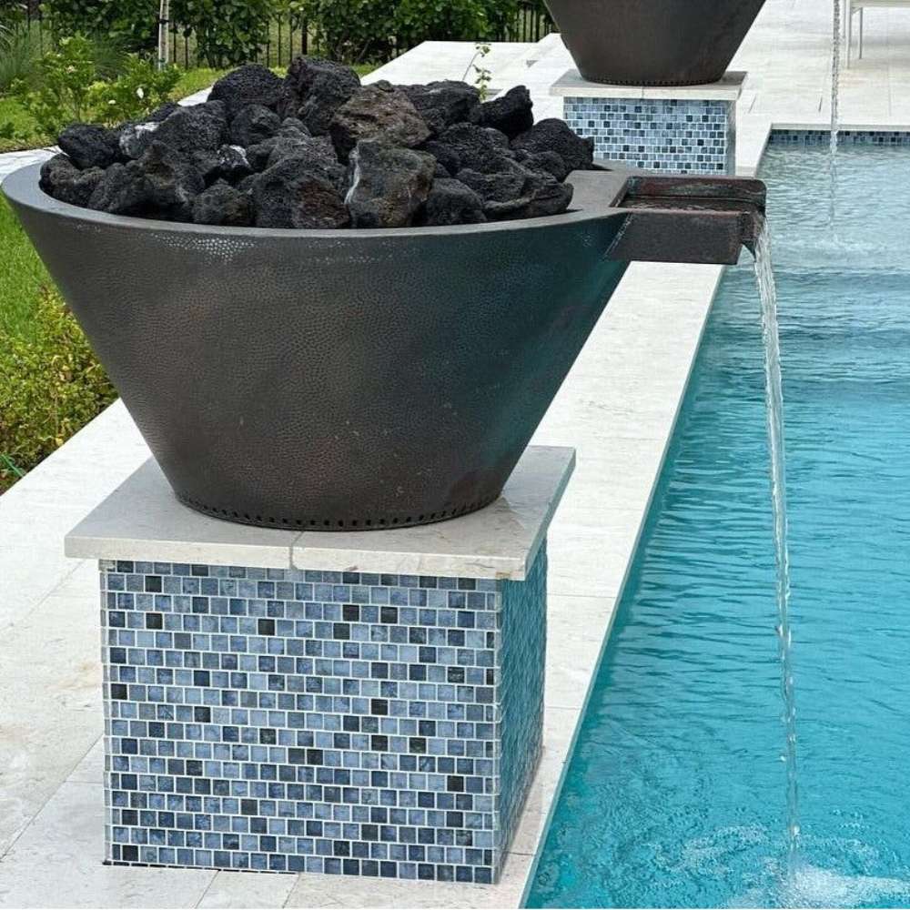 Round gas fire and water bowl on tiled pedestal with dark rocks; water pouring to the pool