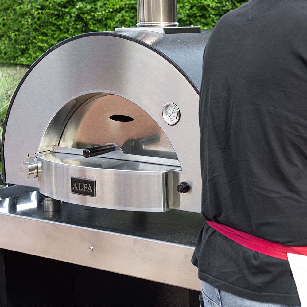 Stainless gas pizza oven with arched dome, front burner, and control knob; cover nearby.
